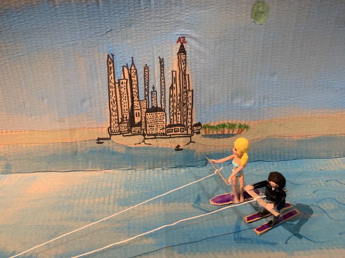 Deux figurines sur une planche de wakeboard tirée par un bateau en plastique sur une surface bleue.