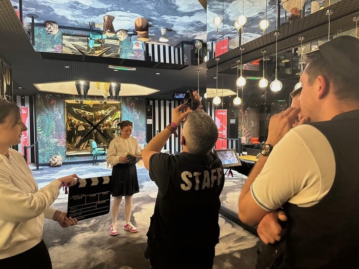 Collaboratrice jouant devant la caméra du coach, clap tenu par sa collègue, pendant un team building créatif